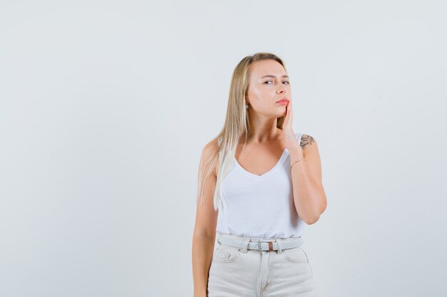 Dama rubia sosteniendo la mano cerca de la boca en camiseta, pantalones y mirando vacilante