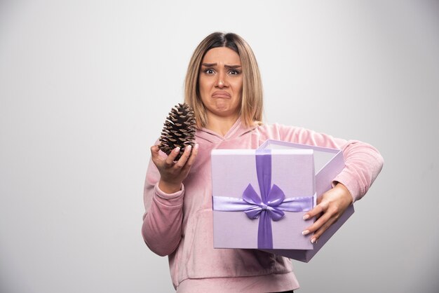 La dama rubia con sudadera rosa saca un cono de roble de la caja de regalo y se siente insatisfecha.