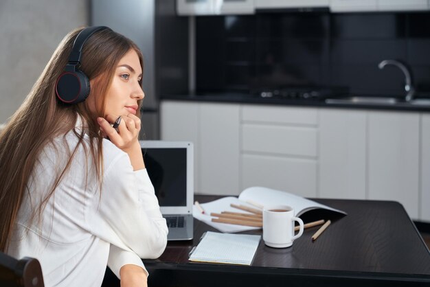 Dama soñadora en auriculares sentada en la mesa con una computadora portátil