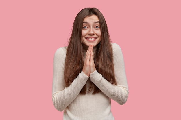 La dama sonriente complacida tiene una expresión suplicante, mantiene las palmas juntas, reza por todo lo bueno, tiene el cabello largo y oscuro, usa ropa informal