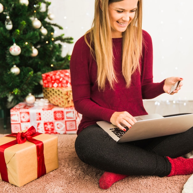 Dama sonriente con laptop y tarjeta de plástico cerca de cajas de regalo y árbol de navidad