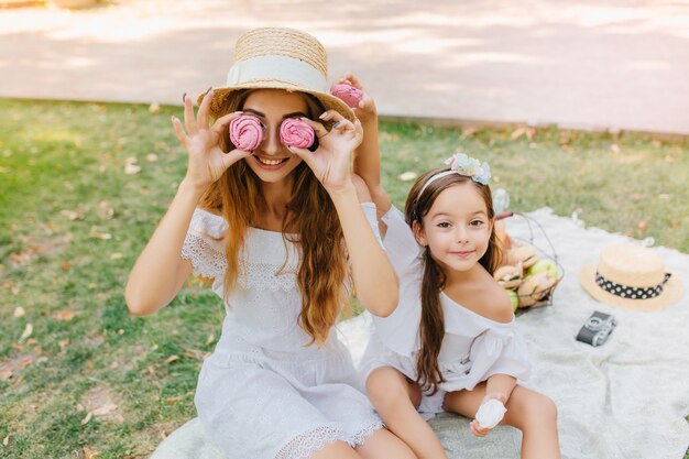 Dama sonriente con vestido blanco sosteniendo pan de jengibre rosa como gafas, sentada sobre una manta con mi hija. Niña bonita con cinta posando junto a la madre en broma durante el picnic.