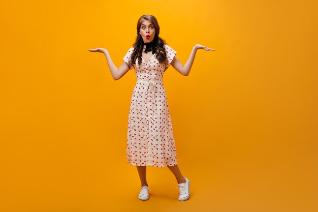 Dama sorprendida encogiéndose de hombros sobre fondo naranja. Moda joven con cabello ondulado en vestido largo blanco mirando a cámara.