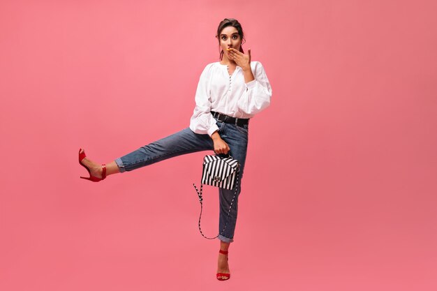 La dama sorprendida levanta la pierna y posa con un elegante bolso sobre fondo rosa. Chica divertida en blusa de manga larga y tacones rojos mira a la cámara. .