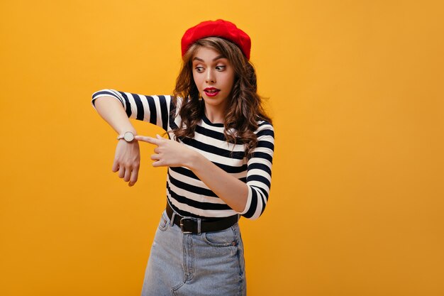 Dama sorprendida en puntos de boina roja para mirar. Linda mujer joven con labios brillantes y bonito sombrero posando sobre fondo naranja.