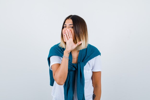 Dama con suéter atado en camiseta blanca contando secretos detrás de la mano y mirando curiosa, vista frontal.