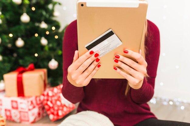 Dama con tableta y tarjeta de plástico cerca de cajas de regalo y árbol de navidad