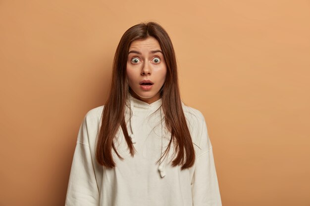 Dama temerosa aguanta la respiración por el shock, tiene los ojos salidos, tiene el pelo largo, está vestida con una sudadera blanca, tiene maquillaje natural