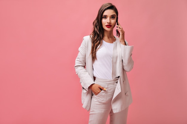 La dama de traje blanco mira a la cámara y habla por teléfono. Chica moderna en camiseta ligera y chaqueta color crema posa sobre fondo aislado.