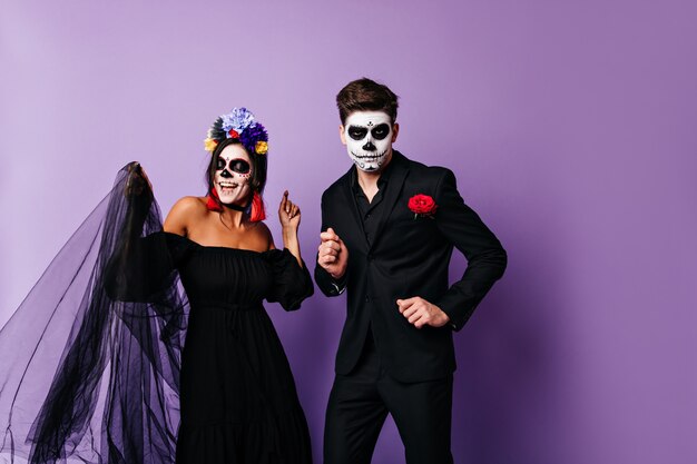 Dama traviesa de negro y su novio serio bailan sobre fondo morado. Retrato de pareja en trajes de halloween de estilo mexicano.