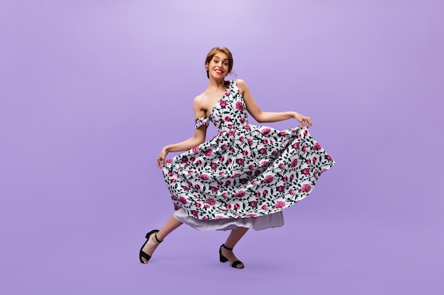 Dama con vestido estampado floral hace reverencia. Sonriente mujer de moda en ropa de verano brillante moderno posando sobre fondo aislado.