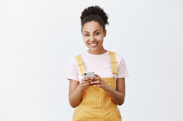 Dame tu número. Retrato de encantadora mujer joven de piel oscura coqueta y femenina con un mono amarillo, sosteniendo un teléfono inteligente y escribiendo el correo electrónico de un amigo, mirando con una hermosa sonrisa