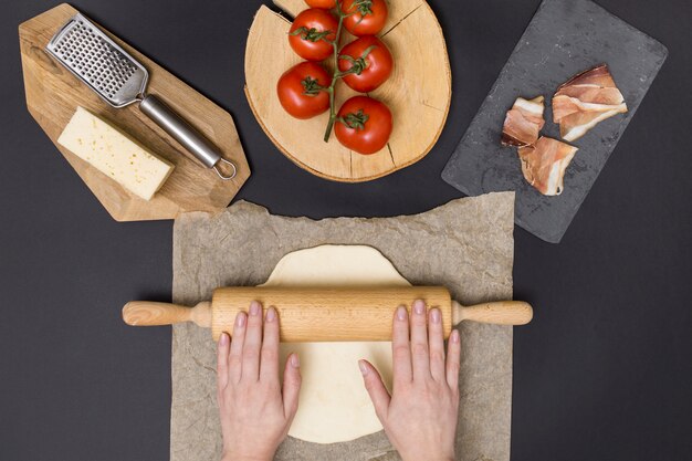 Dé la masa de la pizza del balanceo en el papel de pergamino con el ingrediente de la pizza en fondo negro