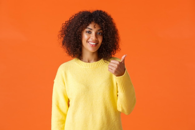 Deberías venir a verlo. Suéter amarillo despreocupado atractivo de la muchacha del inconformista, invitando a entrar apuntando el pulgar detrás de ella y sonriendo, recomendar el producto o el servicio de la empresa, fondo naranja