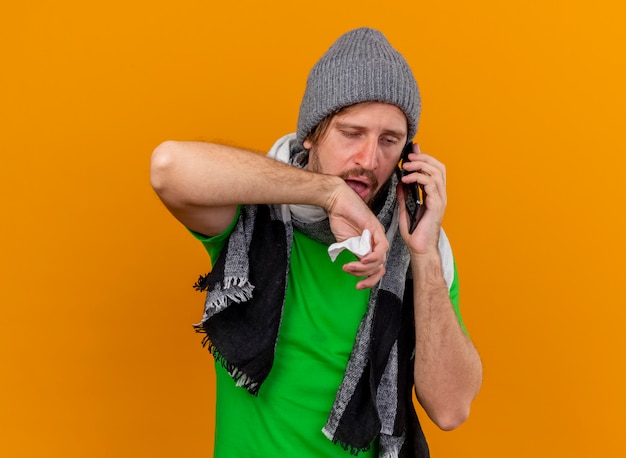 Débil joven apuesto enfermo eslavo con sombrero de invierno y bufanda hablando por teléfono sosteniendo una servilleta mirando recto manteniendo la mano en el aire aislado sobre fondo naranja con espacio de copia