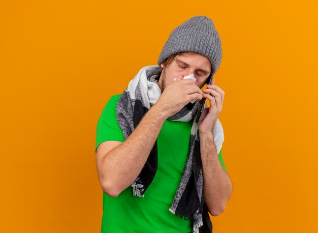 Débil joven apuesto enfermo con gorro de invierno y bufanda hablando por teléfono sosteniendo una servilleta limpiando la nariz con los ojos cerrados aislados en la pared naranja