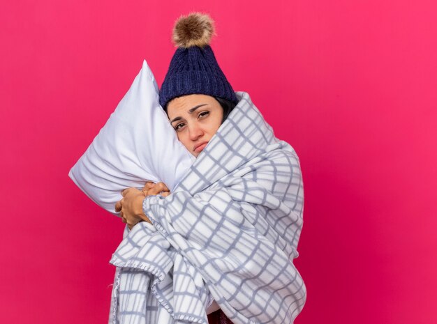 Débil joven caucásica enferma con gorro de invierno y bufanda envuelta en una almohada a cuadros abrazando poniendo la cabeza sobre ella mirando a cámara aislada sobre fondo carmesí con espacio de copia