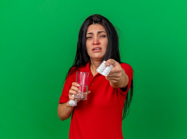 Débil joven caucásica enferma sosteniendo paquete de tabletas vaso de agua y servilleta estirando paquete de tabletas hacia la cámara mirando a cámara aislada sobre fondo verde con espacio de copia