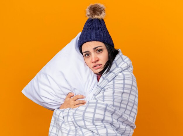 Débil joven caucásica enferma vestida con gorro de invierno y bufanda envuelta en una almohada a cuadros abrazando mirando a cámara aislada sobre fondo naranja con espacio de copia