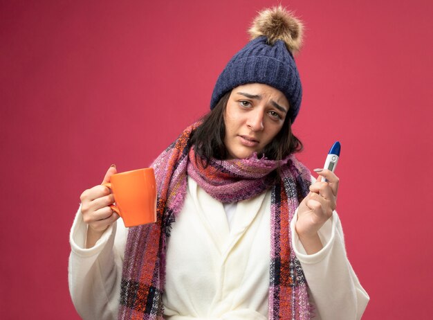 Débil joven caucásica enferma vistiendo bata gorro de invierno y bufanda sosteniendo una taza de té y termómetro mirando a cámara aislada sobre fondo carmesí