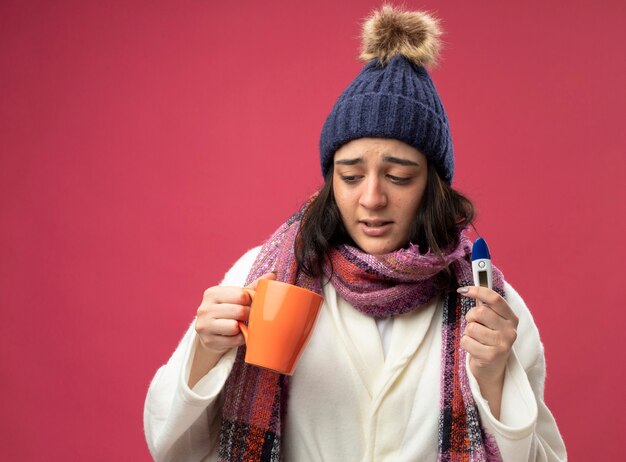 Débil joven caucásica enferma vistiendo bata gorro de invierno y bufanda sosteniendo una taza de té y un termómetro mirando dentro de la taza aislada en la pared carmesí con espacio de copia