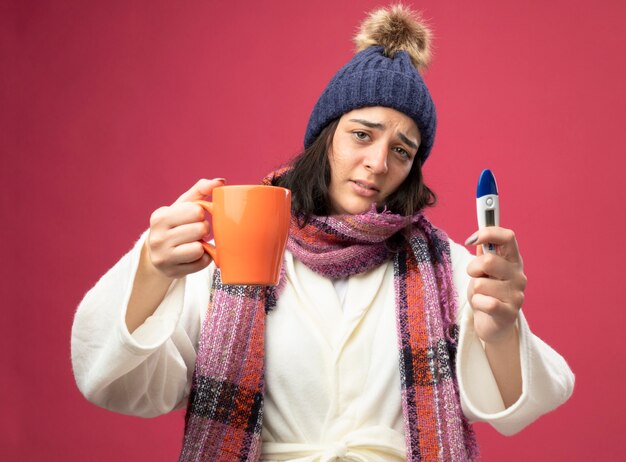 Débil joven caucásica enferma vistiendo bata sombrero de invierno y bufanda estirando una taza de té y sosteniendo el termómetro aislado en la pared carmesí