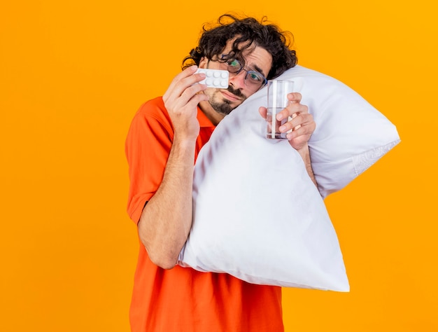 Débil joven caucásico enfermo con gafas abrazando la almohada tocando la cara con ella sosteniendo un paquete de tabletas médicas y un vaso de agua mirando tabletas aisladas sobre fondo naranja con espacio de copia