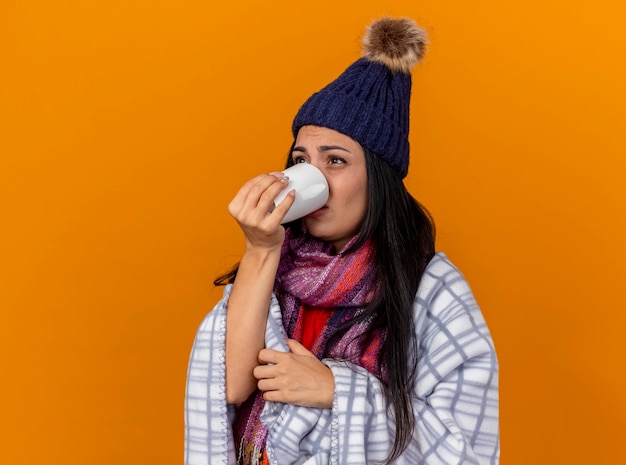 Débil joven enferma con gorro de invierno y bufanda envuelta en cuadros de pie en la vista de perfil bebiendo una taza de té mirando directamente aislado en la pared naranja