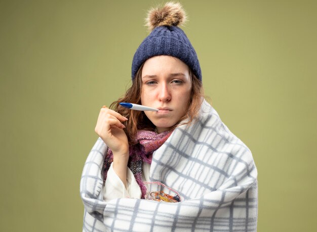 Débil joven enferma vestida con túnica blanca y gorro de invierno con bufanda envuelta en cuadros poniendo termómetro en la boca aislado en verde oliva