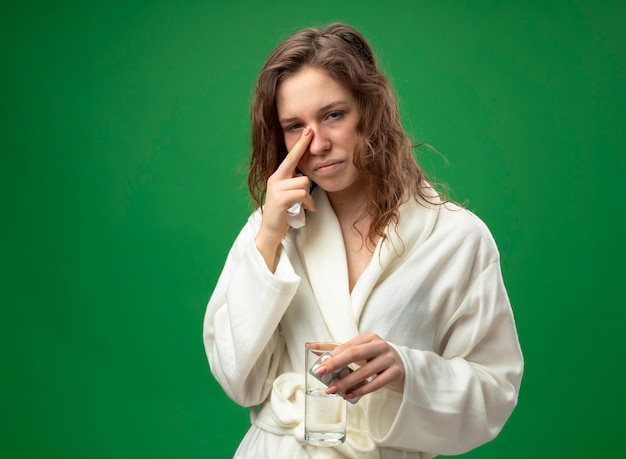 Débil joven enferma vestida con túnica blanca sosteniendo un vaso de agua con pastillas poniendo el dedo en la nariz aislado en verde con espacio de copia