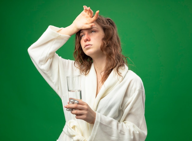 Foto gratuita débil joven enferma vestida con túnica blanca sosteniendo un vaso de agua con pastillas y poniendo la mano en la frente aislada en verde