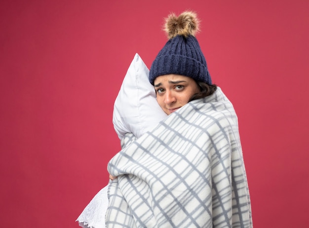 Débil joven enferma vistiendo bata gorro de invierno y bufanda envuelta en cuadros de pie en la vista de perfil abrazando la almohada mirando al frente aislado en la pared rosa