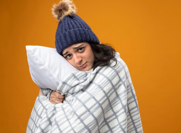 Débil joven enferma vistiendo bata gorro de invierno envuelto en cuadros de pie en la vista de perfil abrazando la almohada mirando al frente aislado en la pared naranja