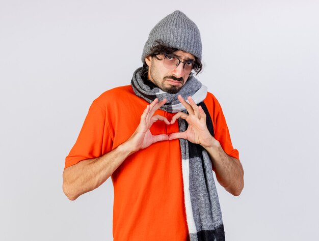 Débil joven enfermo con gafas, gorro y bufanda de invierno mirando al frente haciendo signo de corazón aislado en la pared blanca