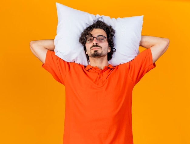 Débil joven enfermo con gafas sosteniendo la almohada debajo de la cabeza mirando al frente aislado en la pared naranja