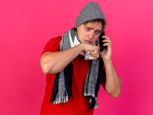 Débil joven guapo rubio enfermo con sombrero de invierno y bufanda hablando por teléfono sosteniendo la servilleta manteniendo la mano cerca de la boca mirando hacia abajo aislado sobre fondo carmesí con espacio de copia