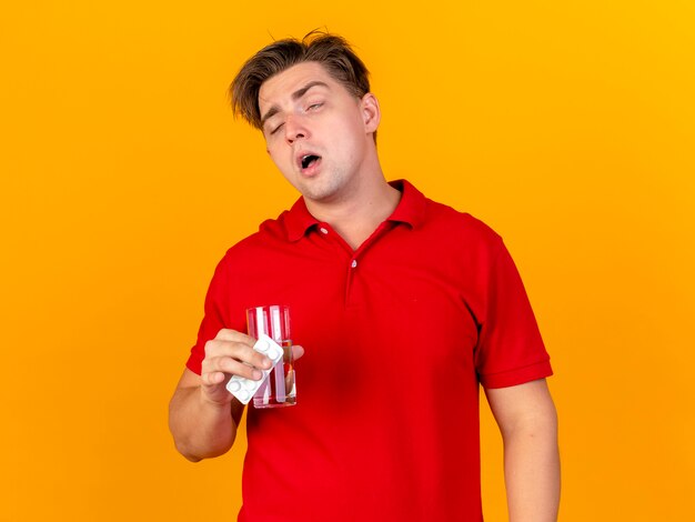 Débil joven guapo rubio enfermo sosteniendo un paquete de tabletas médicas y un vaso de agua aislado sobre fondo naranja