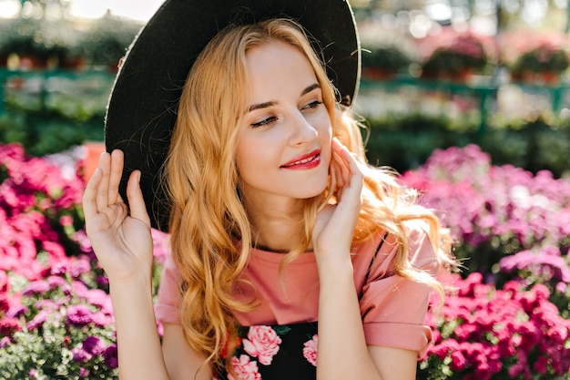 Debinair mujer caucásica posando en frint de flores rosadas. Mujer elegante pensativa con sombrero disfrutando de día de verano.