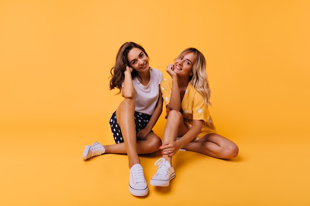 Debonair bronceó a chicas con zapatos deportivos blancos posando en el suelo. Hermanas relajadas de buen humor relajándose juntas.