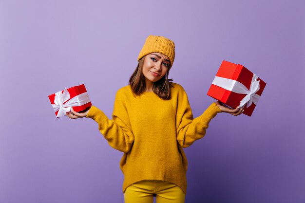 Debonair chica caucásica sosteniendo presnets de cumpleaños con sonrisa triste. Retrato de interior de adorable joven en traje de punto amarillo de pie sobre morado.