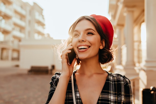 Debonair mujer caucásica en boina de moda hablando por teléfono. Foto al aire libre de la romántica chica francesa con sonrisa alegre.