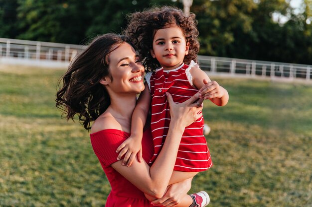 Debonair mujer europea disfrutando de la mañana del fin de semana con su pequeña hija. Hermosa joven mamá posando en el parque con niño rizado.