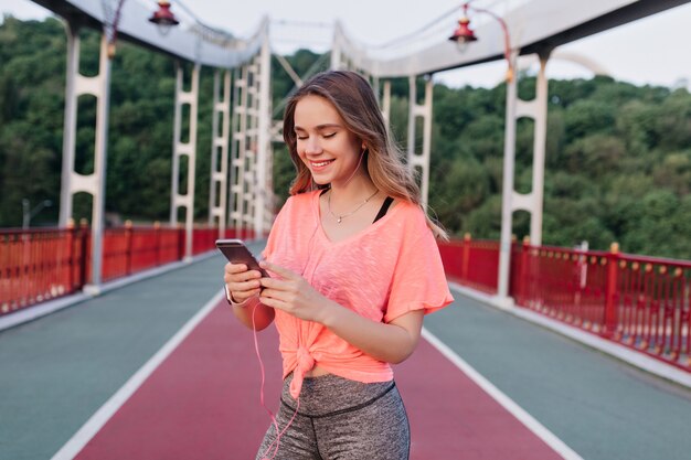 Debonair mujer rubia con teléfono durante el entrenamiento. Niña sonriente en traje casual posando en el estadio por la mañana.