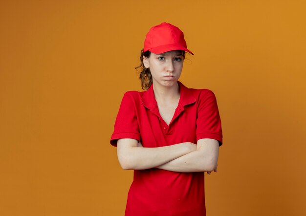 Decepcionada joven bastante repartidora en uniforme rojo y gorra de pie con postura cerrada aislada sobre fondo naranja con espacio de copia