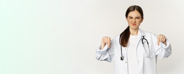 Foto gratuita decepcionada joven doctora doctora señalando con el dedo hacia abajo y haciendo una mueca con disgusto desaprobación
