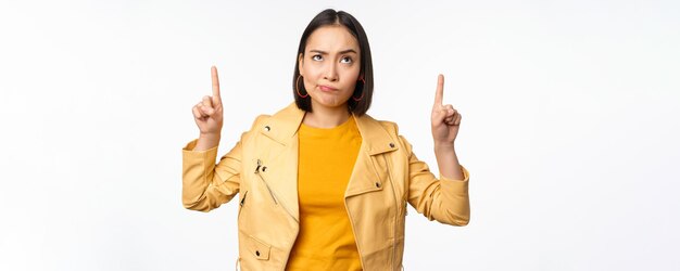 Decepcionada modelo femenina coreana frunciendo el ceño y haciendo una mueca escéptica apuntando hacia arriba y mirando molesta a la pancarta en la parte superior de pie sobre fondo blanco.