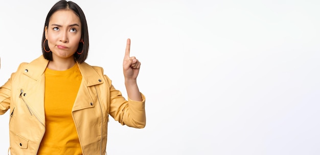 Decepcionada modelo femenina coreana frunciendo el ceño y haciendo una mueca escéptica apuntando hacia arriba y mirando molesta a la pancarta en la parte superior de pie sobre fondo blanco.