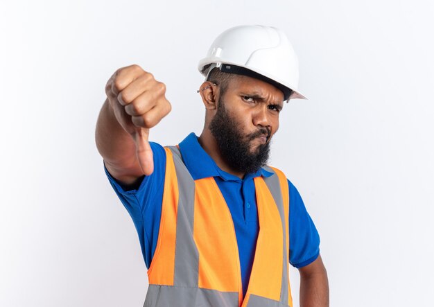 Decepcionado joven constructor afroamericano en uniforme con casco de seguridad pulgar hacia abajo aislado sobre fondo blanco con espacio de copia