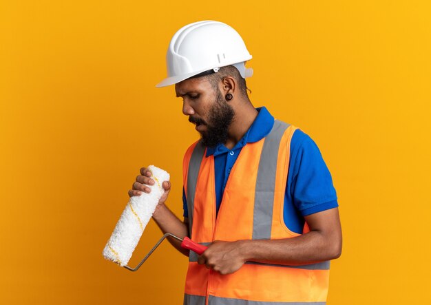 Decepcionado joven constructor afroamericano en uniforme con casco de seguridad sosteniendo y mirando el rodillo de pintura aislado sobre fondo naranja con espacio de copia