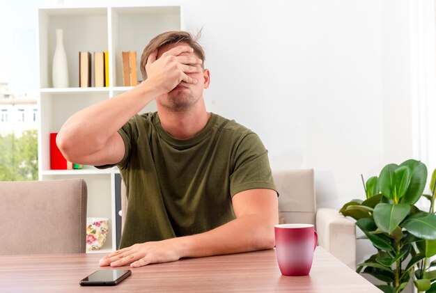 Decepcionado joven guapo rubio se sienta a la mesa con la taza y el teléfono poniendo la mano en la cara cerrando los ojos dentro de la sala de estar